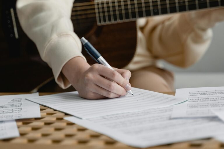 Person writing music on sheet paper with an acoustic guitar in a cozy home setting.